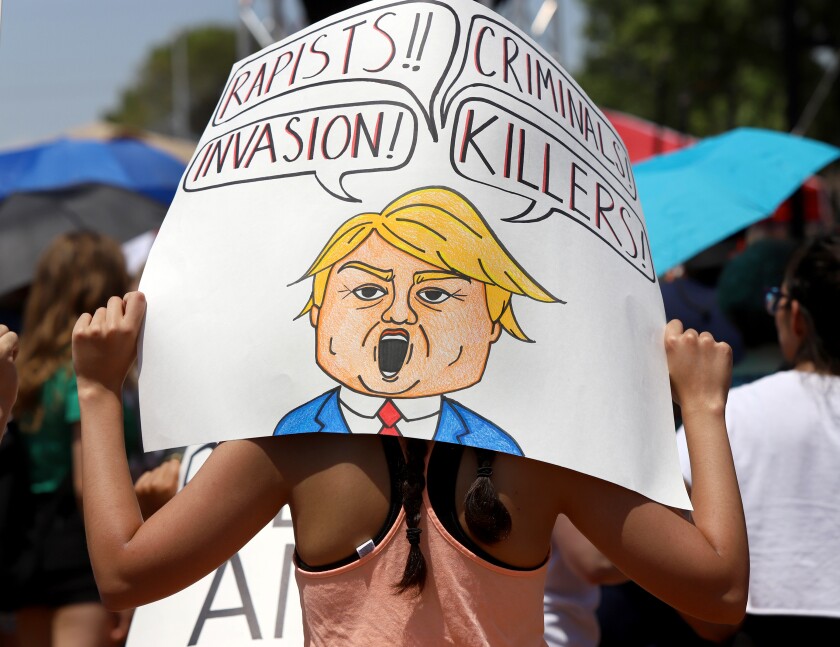 A woman at an El Paso protest holds a sign with a cartoon Trump and speech bubbles that read "Rapists!!" "Criminals!" "Invasion!" "Killers!"