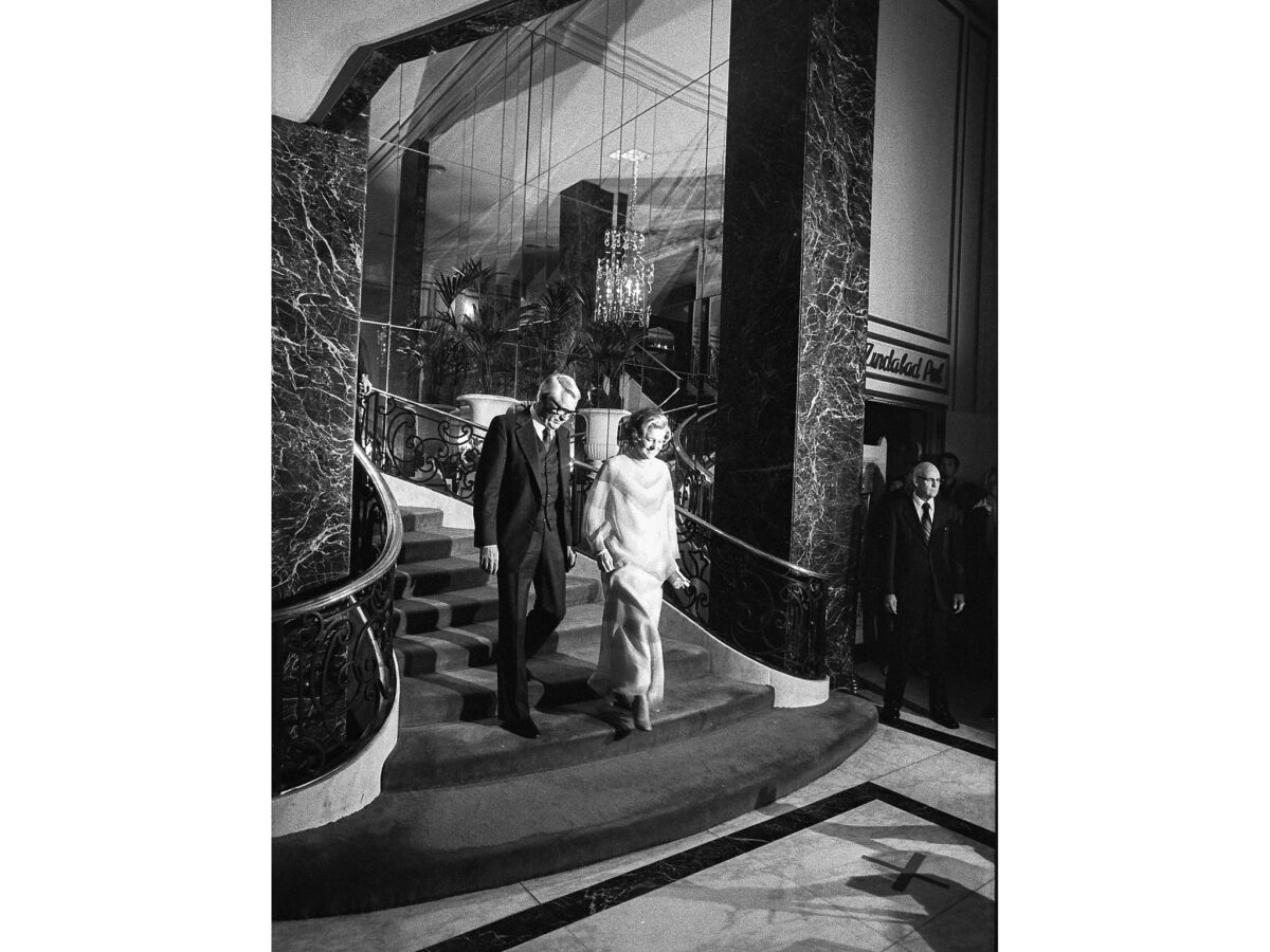 A man and a woman descend a grand mirrored staircase flanked by marble columns.