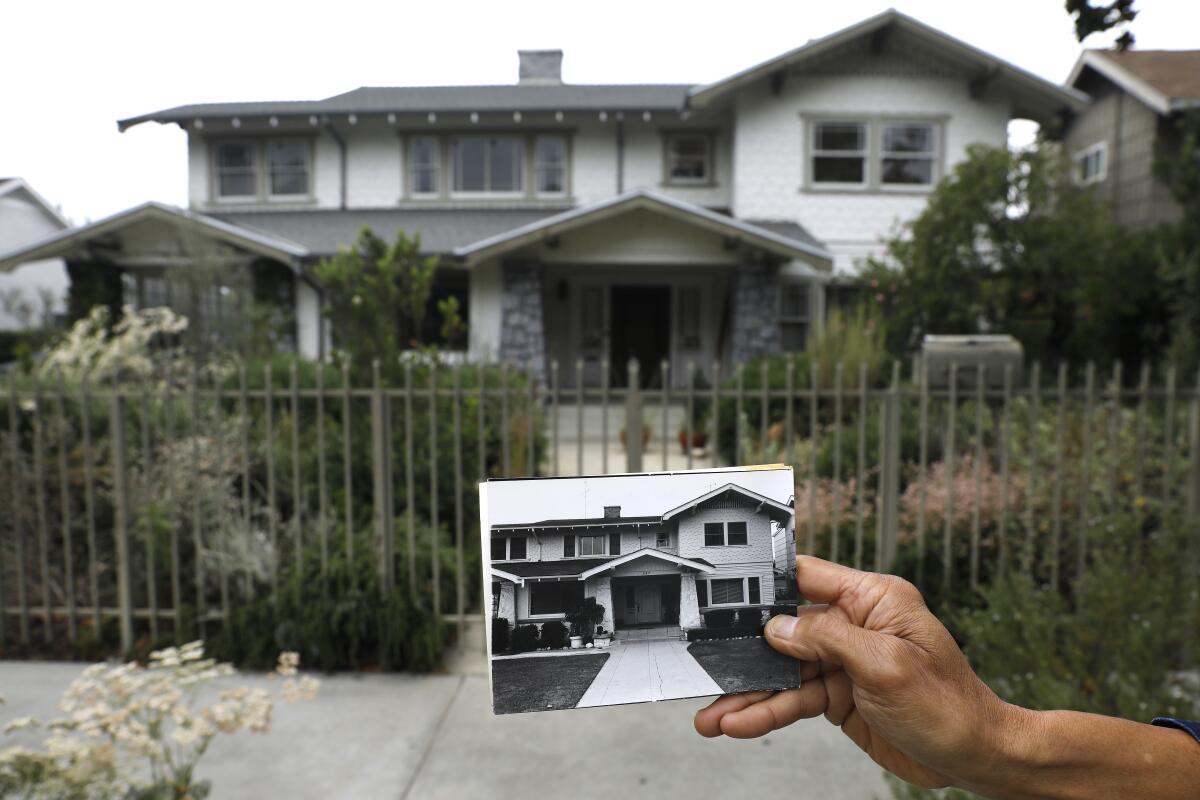 A man's hand holds up an old photo of a house in front of the same house today