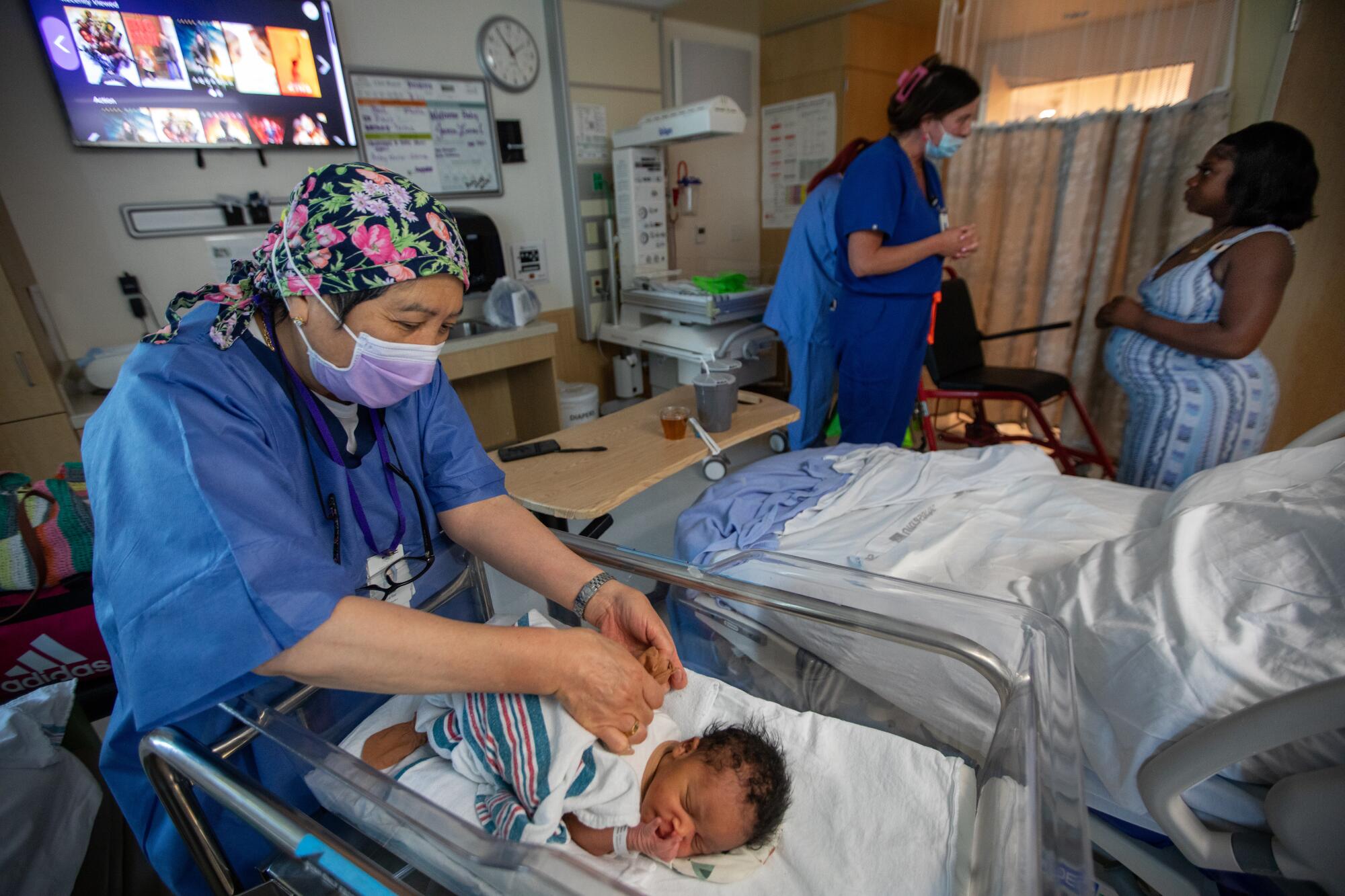 A nurse wraps a baby in a blanket as another speaks to a woman in the background.
