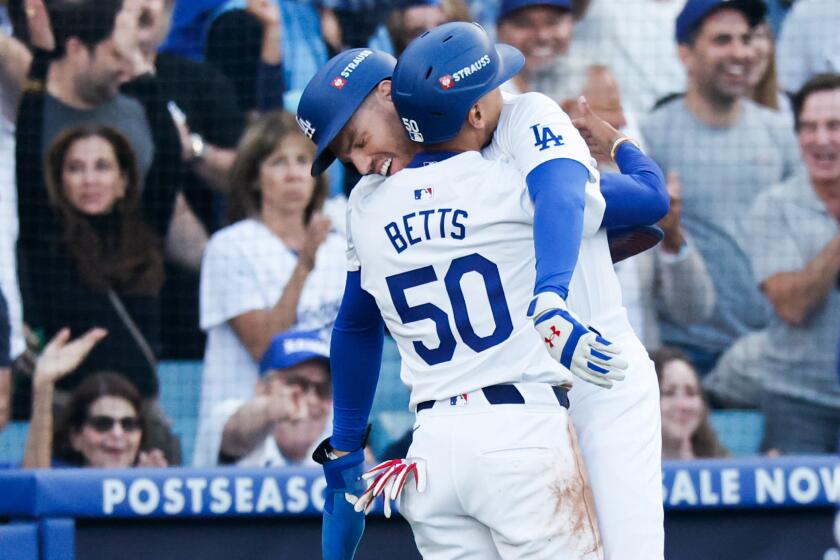 Dodger Mookie Betts hugs teammate Freddie Freeman, helping injured Freeman come to a stop after crossing home plate.