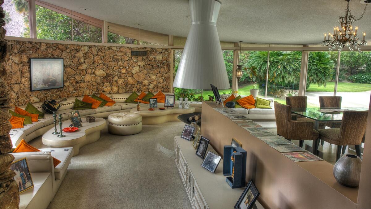 A living and dining area with '60s furniture, chandelier, a curved stone wall, and a glass wall overlooking greenery.