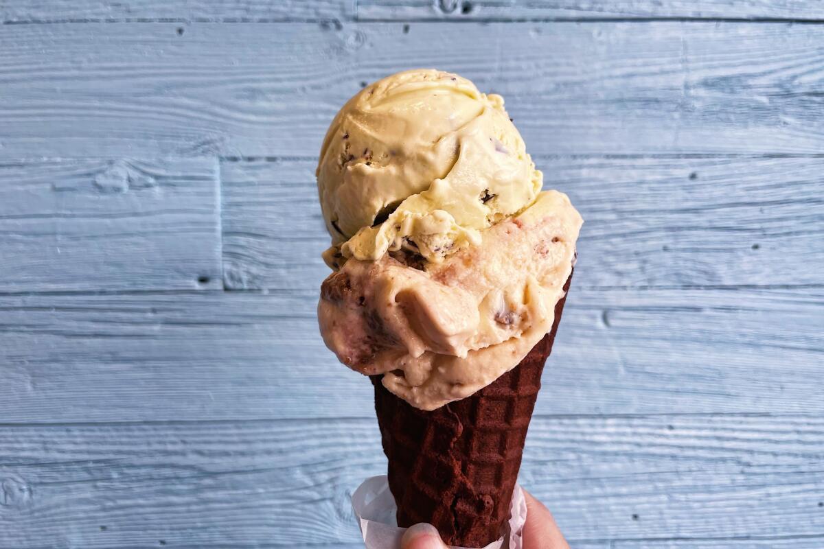 A hand holds a double-scoop chocolate ice cream cone of mint chip and strawberry against a blue wall in Sweet Rose Creamery