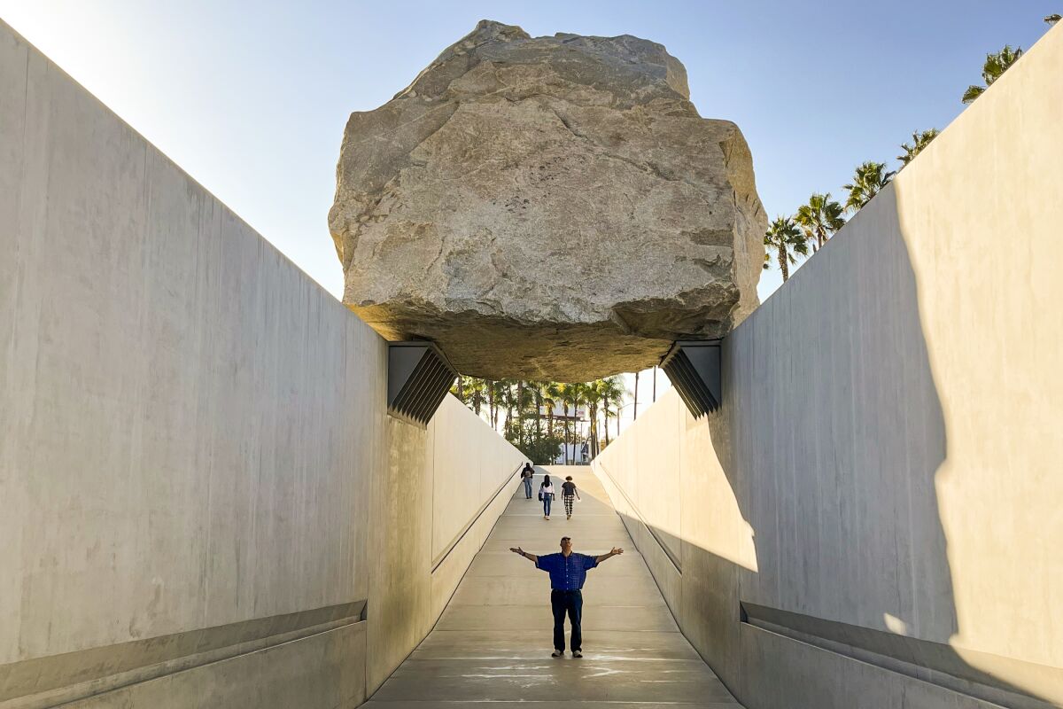 A photograph of a person standing underneath a massive boulder art installation
