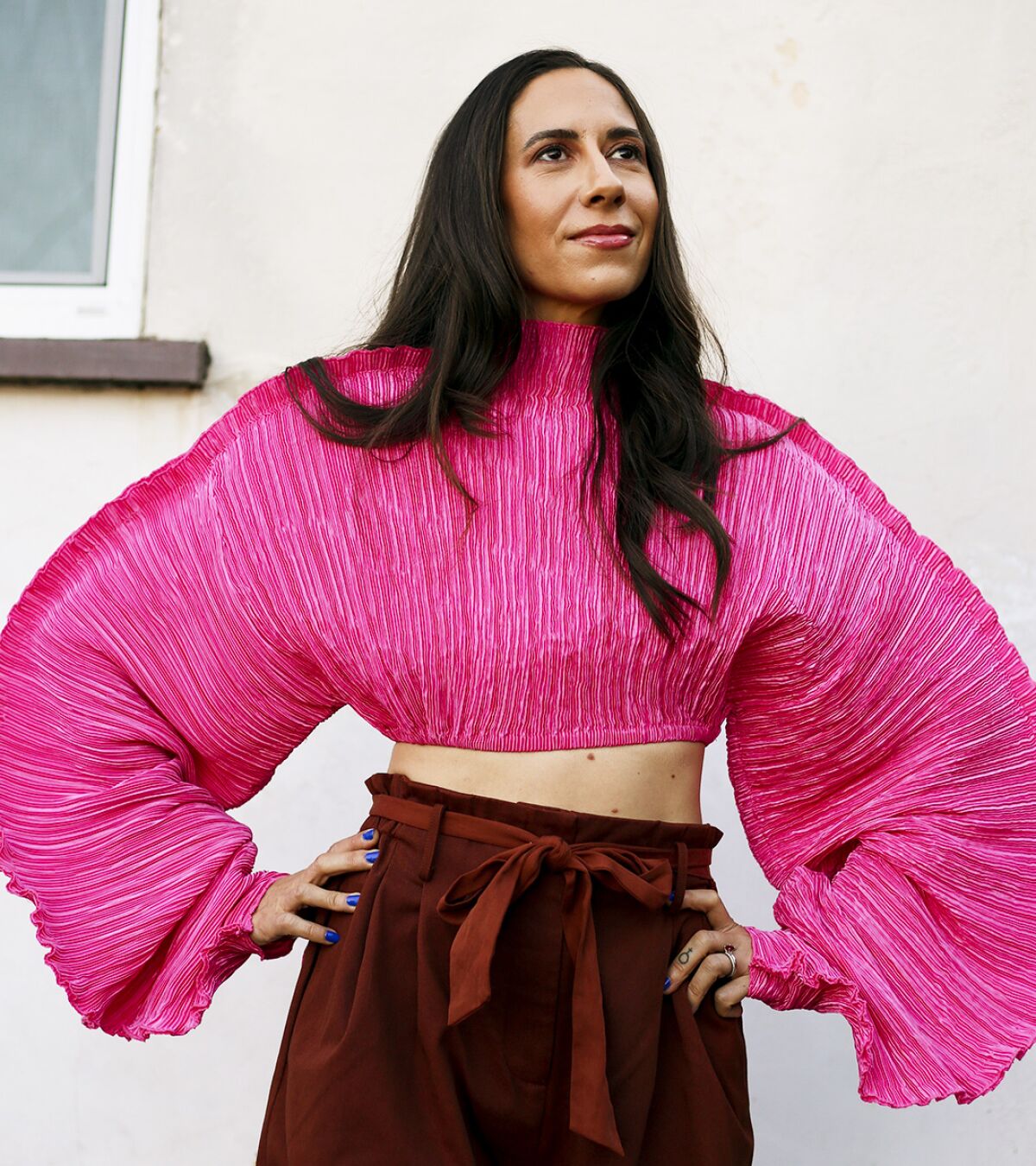 A woman with flowing brunet hair wears a billowy fuschia top and rust-colored bottoms.