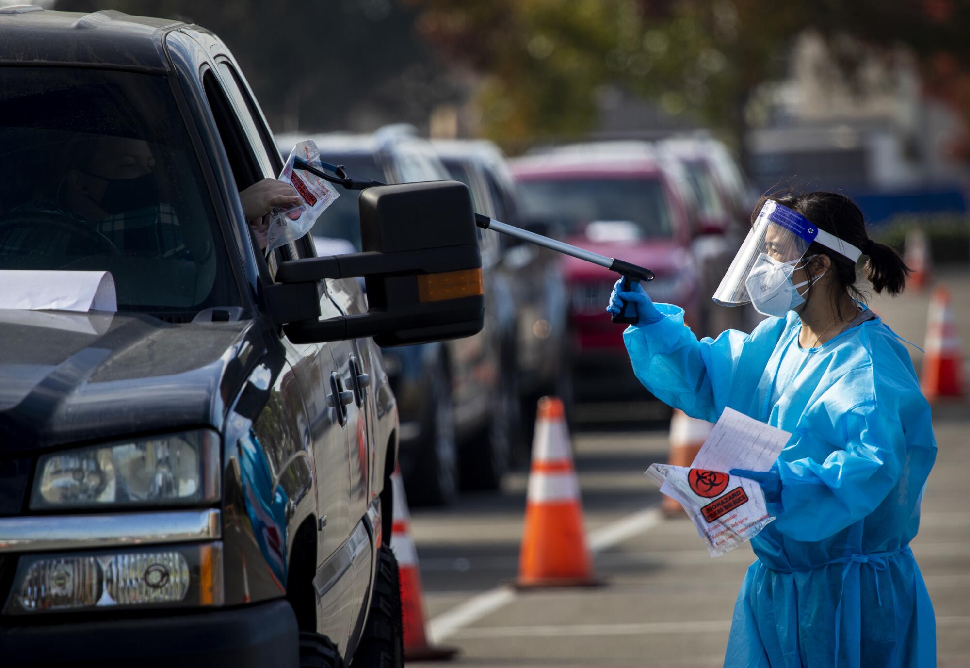 A health worker in protective gear uses a long-handled device to give a motorist a plastic bag with a swab.