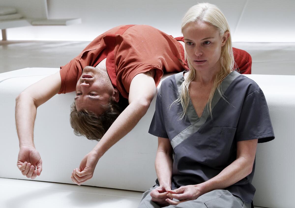 A blond woman sits while a man leans over the back of a couch.