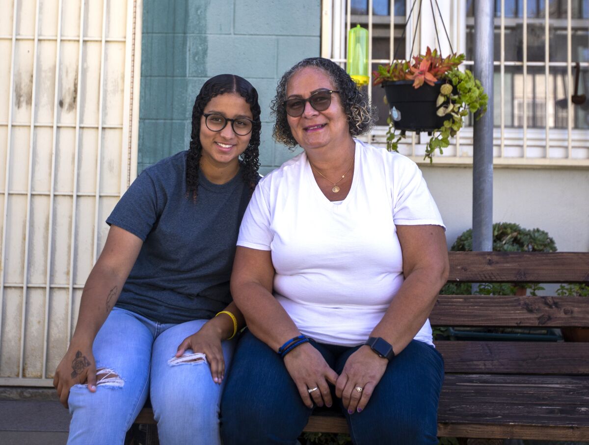 A teenager and her mother sit on a bench outside a home