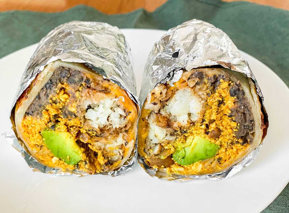 A burrito in foil, halved and open. The insides display avocado, scrambled tofu, tater tots and black beans.
