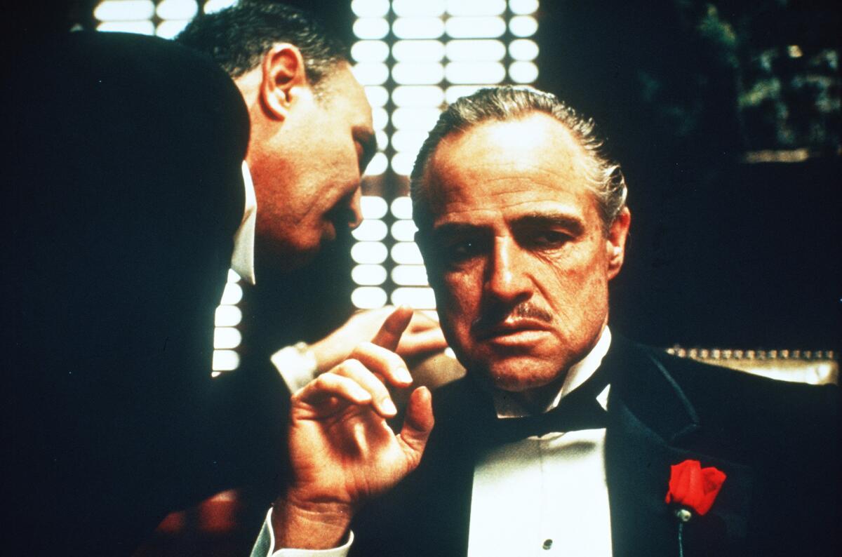 A Mafia don in a tuxedo listens as a man in a tuxedo bends over to speak into his ear.