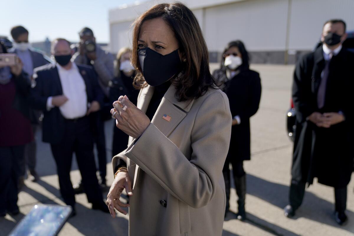 Kamala Harris wearing a mask and tan trench coat with an American flag pin on the lapel