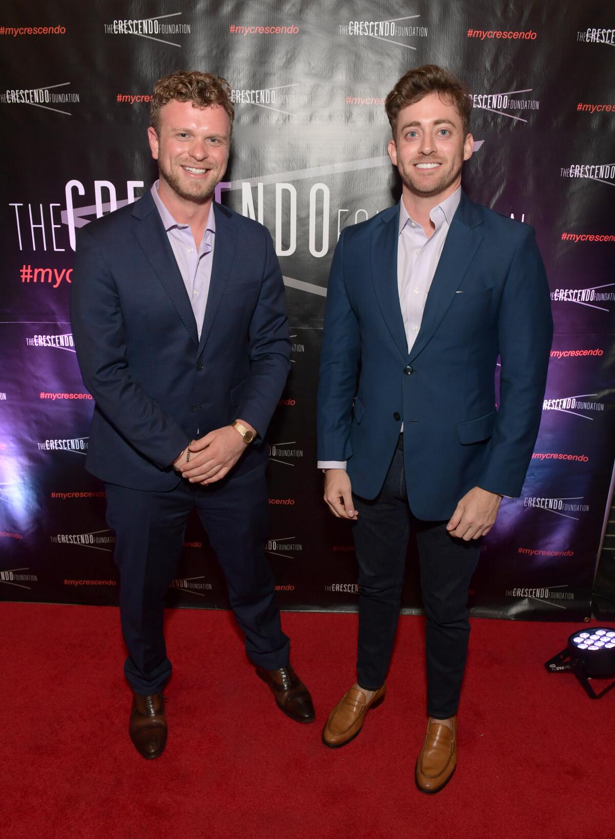 Two men in blue jackets stand on a red carpet.