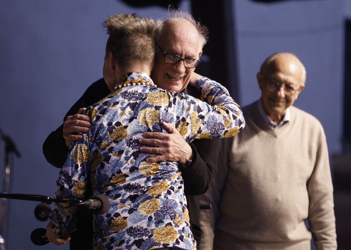 A man wearing a floral shirt hugs another man onstage while a third looks on. 