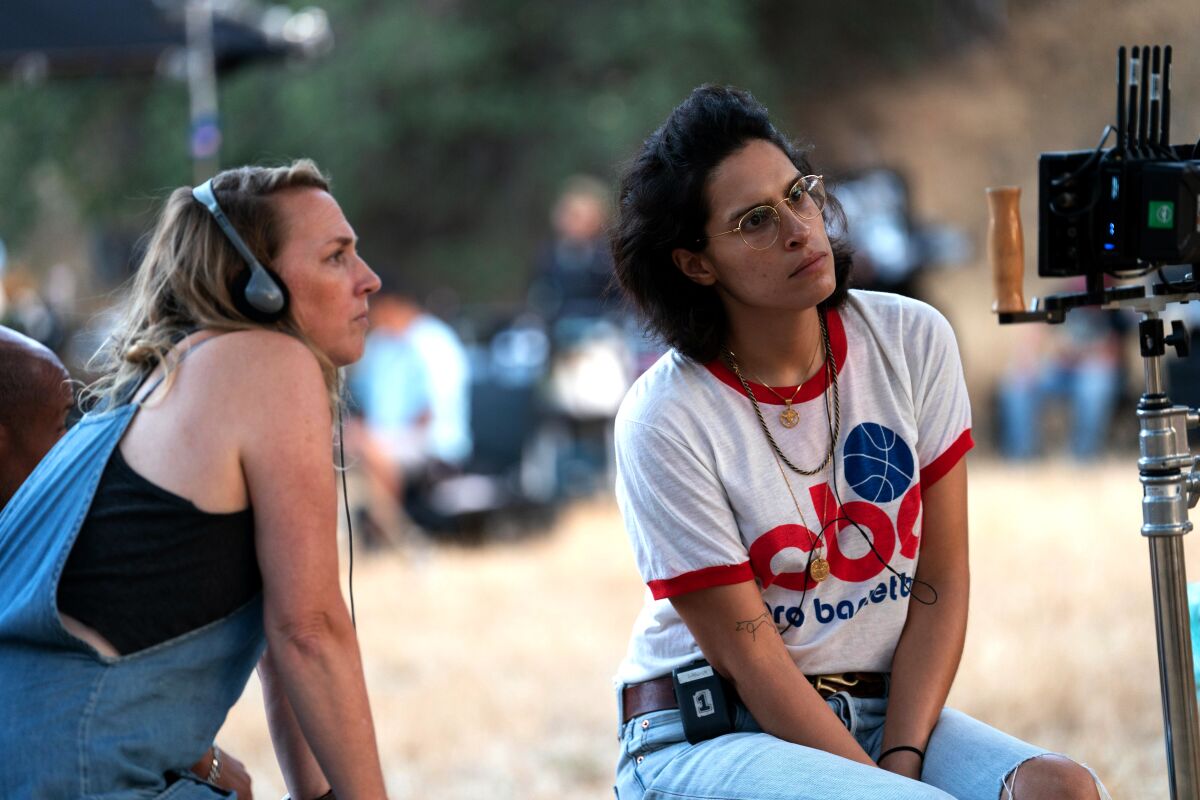 Two women, one wearing headphones, sit on a film set looking at a camera