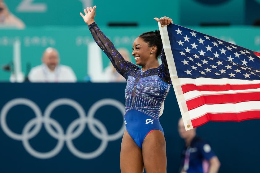 Simone Biles in black and blue sequined bodysuit smiles and waves with U.S. flag in hand at the 2024 Paris Olympics