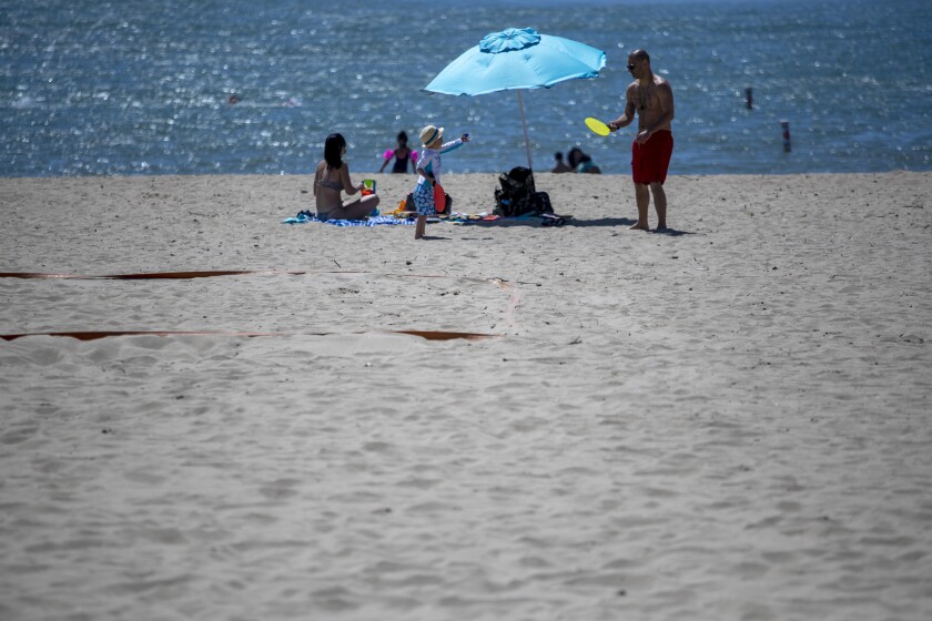 A man throws a Frisbee to a child on the sand as others sit around a beach umbrella