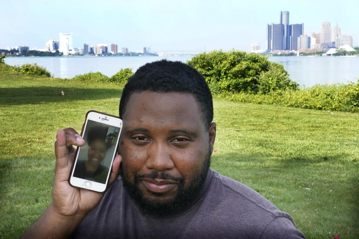 With the cities of Windsor, Ontario, Canada, left, and Detroit, right, seen in the background, Quintin Sweat Jr, poses with his fiancee Renee Harrison, seen on his phone, Tuesday, Aug. 3, 2021, on Belle Isle in Detroit. Sweat and Harrison live only 15 minutes apart by car, with the U.S.-Canada border between them. But the couple, who got engaged in 2019, has only been able to be together three times during the pandemic. (AP Photo/Carlos Osorio)
