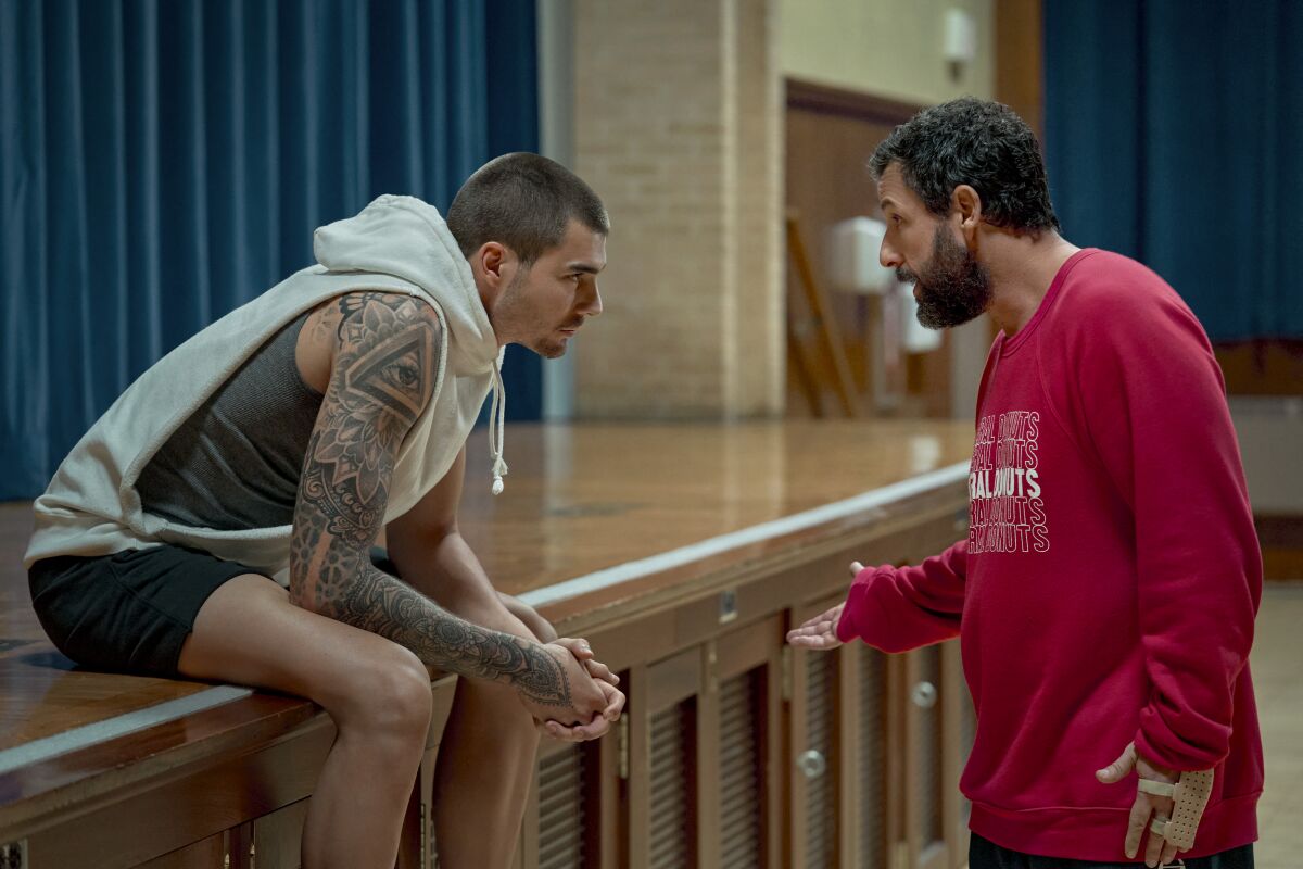 A young man with tattoos and a hoodie sits on the edge of a school auditorium stage; an older bearded man talks to him.