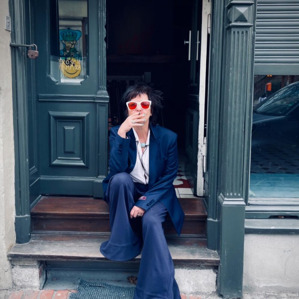 A woman in a blue suit and red shades smokes a cigarette on an urban stoop.