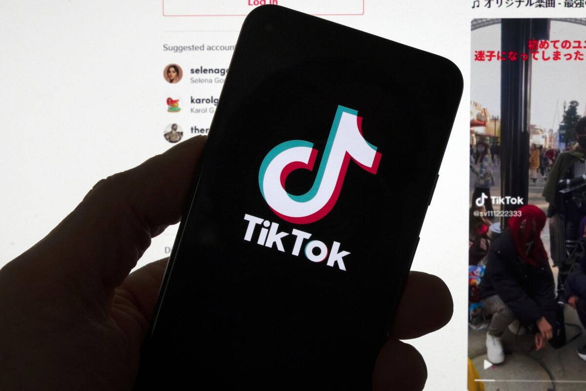 The TikTok logo on a mobile phone in front of a computer displaying the TikTok home page