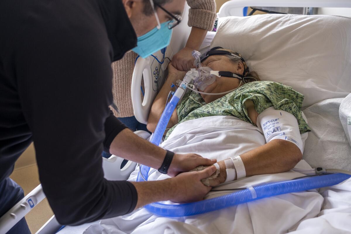A man leans over and holds the hand of a woman attached to a ventilator in a hospital bed