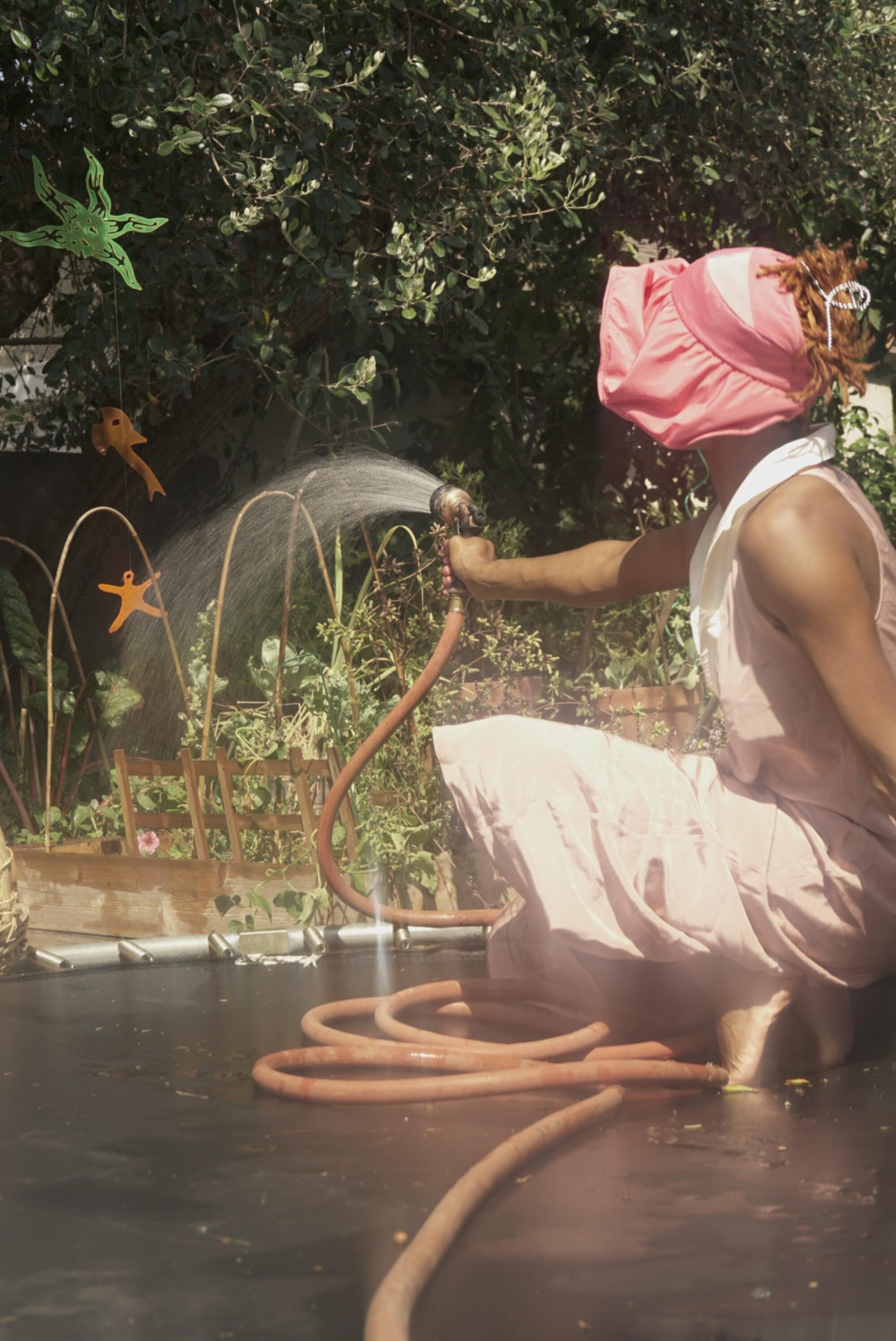 A woman in a pink hat sprays plants with a hose.