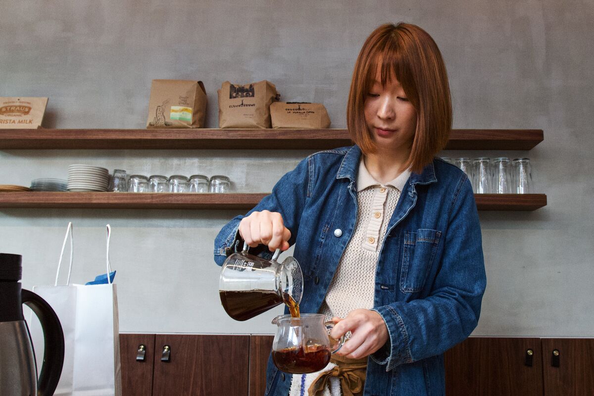 A woman in a coffee shop pours coffee from a glass coffeepot into a glass mug.