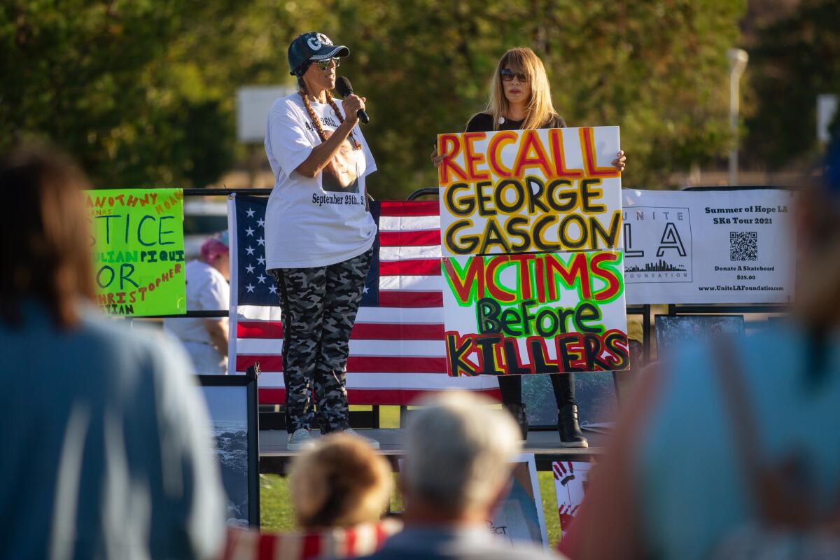 Two women on an outdoor stage, one with a sign that says Recall George Gascon; Victims before killers