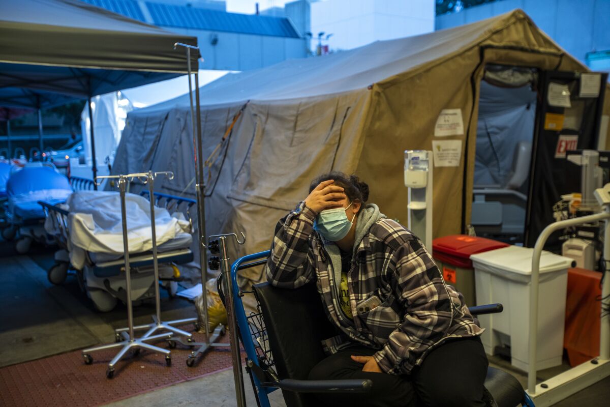 A person in a mask sits in a wheelchair, holding their head, near a tent in a parking lot.