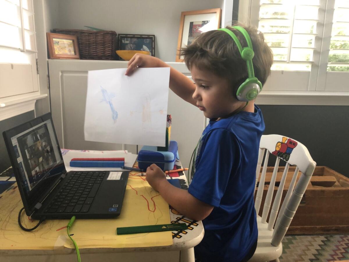 A young boy wearing headphones holds a drawing in front of his laptop camera