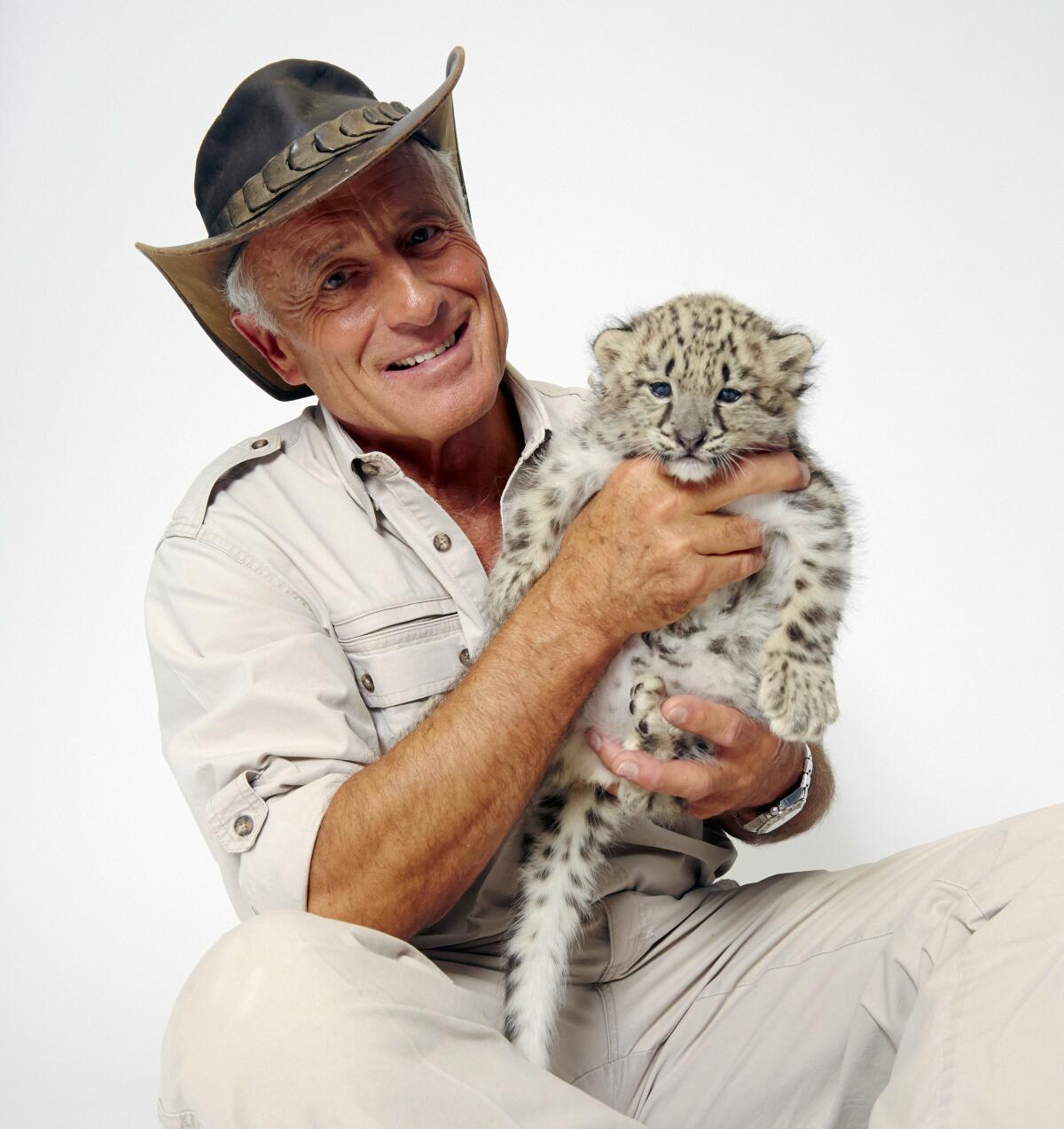 Jack Hanna wears a wide-brimmed hat and holds a snow leopard cub