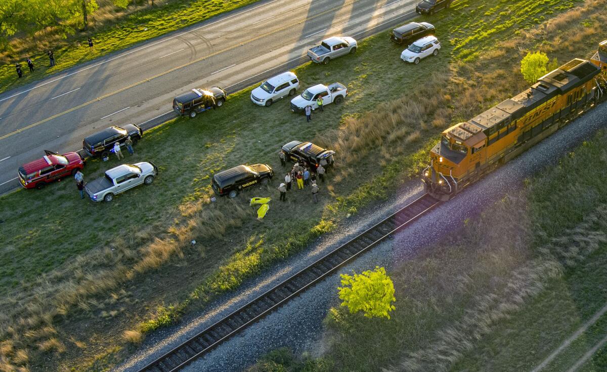 An aerial view of a halted freight train and several vehicles parked in grass next to the tracks.