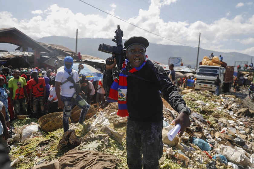 A man in cap and fatigues holds up an assault rifle as he stands amid a pile of trash. A crowd watches from nearby 