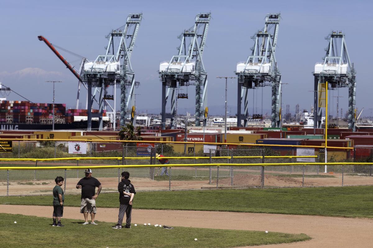 A man coaches two boys on a baseball field while port cranes loom in the background.