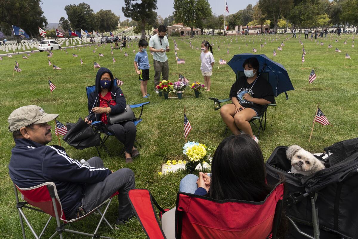 A family sits in folding chairs around a gravestone in a military cemetery with small U.S. flags in the grass