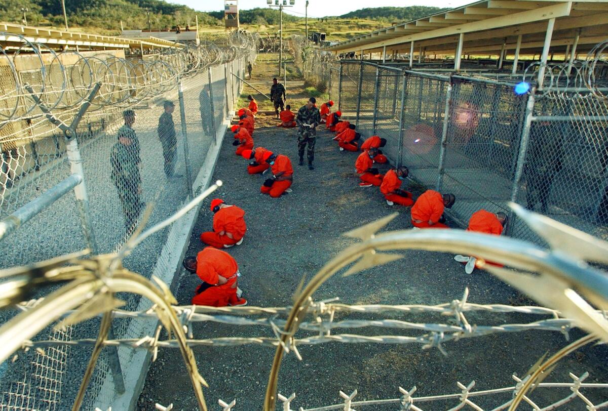 Detainees in orange jumpsuits sit in a holding area under the surveillance of U.S. military police.