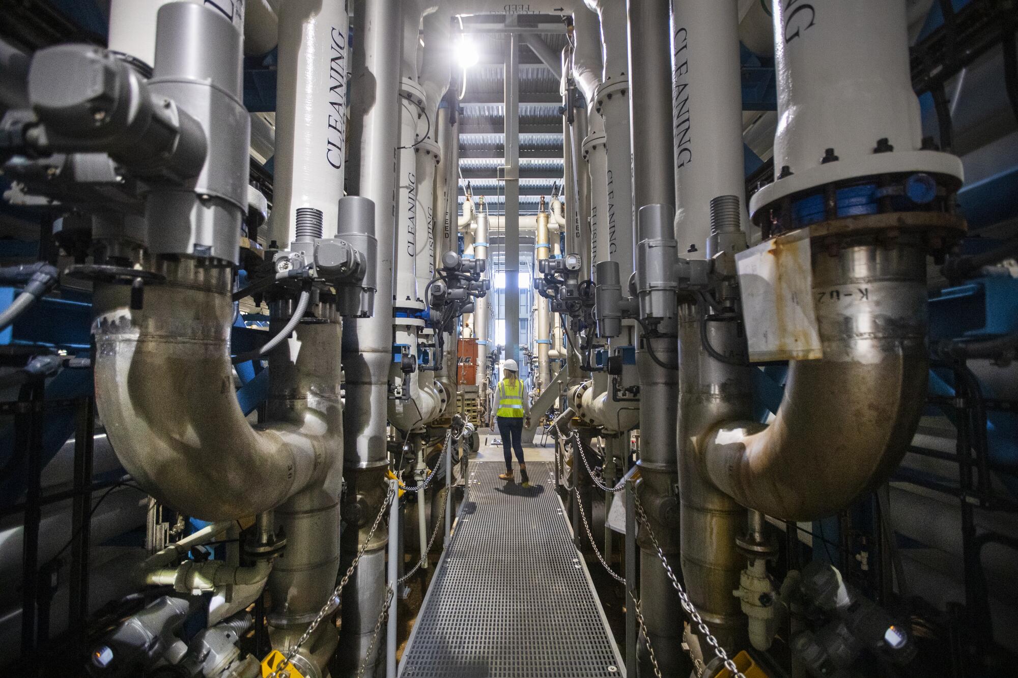 A person in a hardhat walks amid a network of industrial pipes.