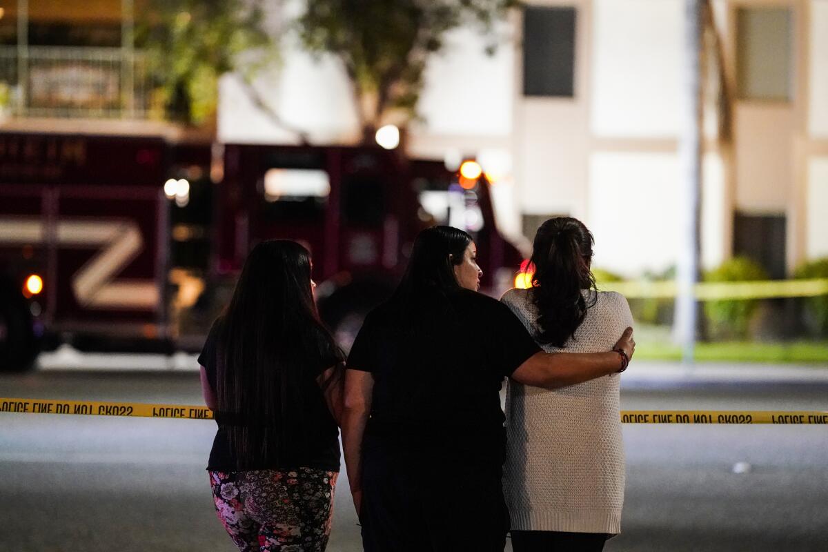 Three women, one with her arm around another, gather across from the scene of a multiple shooting 