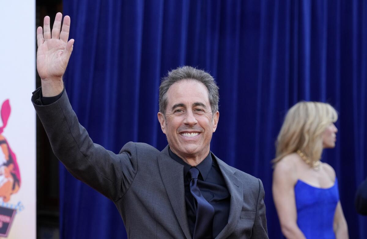 Jerry Seinfeld in a gray suit waving to photographers while standing in front of a blue curtain