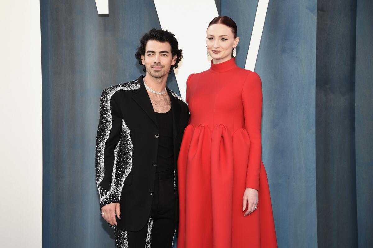 Joe Jonas poses in a sparkly black suit next to Sophie Turner in a red gown against a gray backdrop.