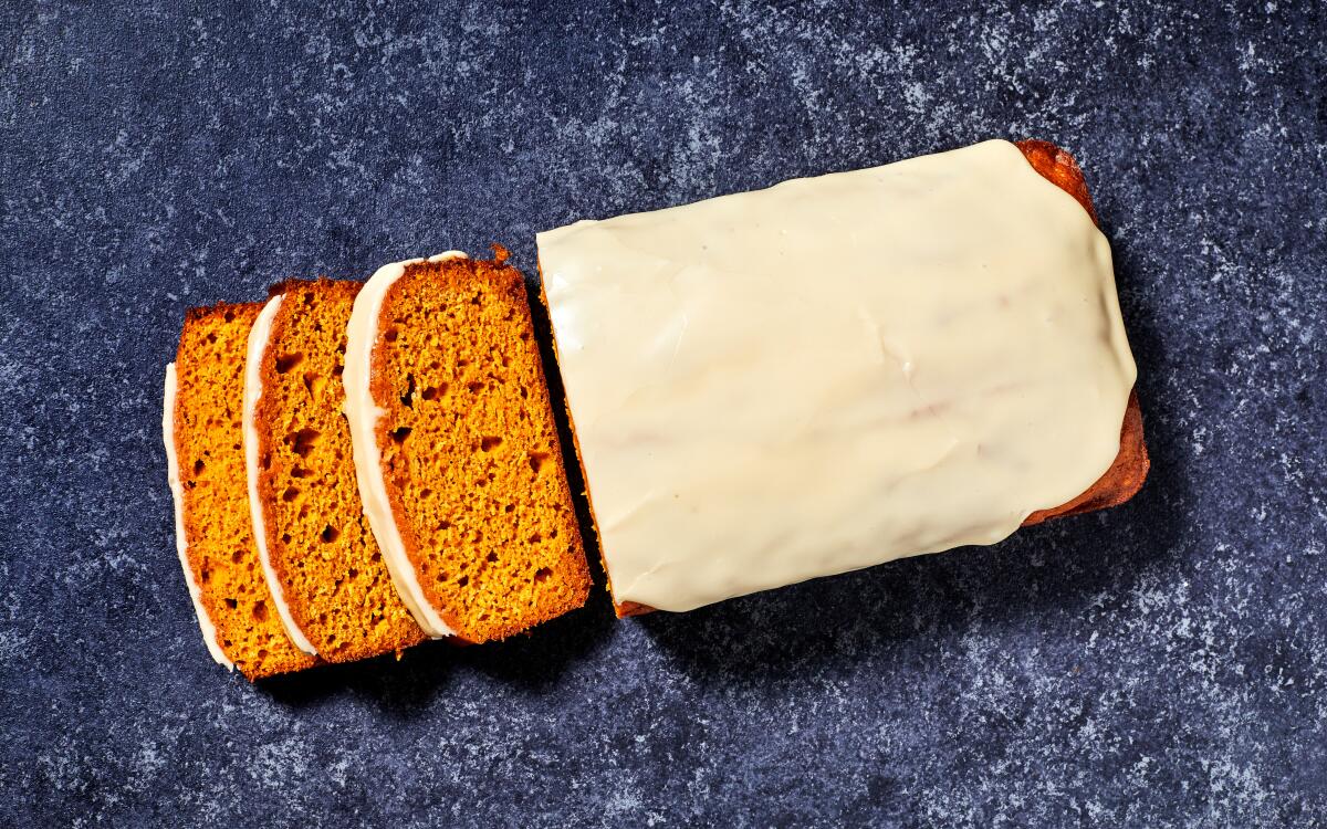 A pumpkin loaf cake topped with white icing has three slices lying next to it.