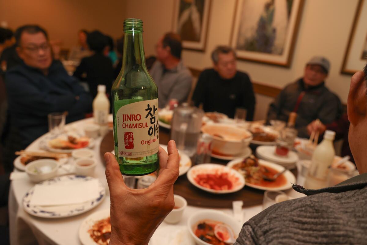 A hand holds up a green glass beverage bottle in a dining room filled with people at round tables.