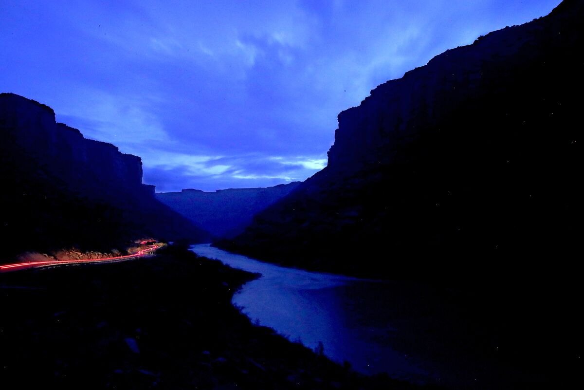 A view of a river and steep hillsides at dusk. A line of red from car taillights is visible along one hillside.
