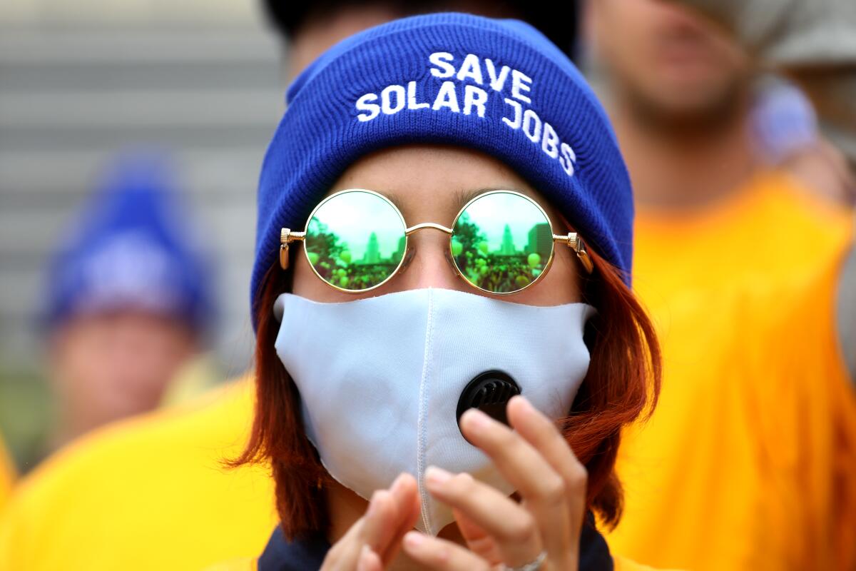 A woman in a face mask and sunglasses wears a beanie that says "Save solar jobs."