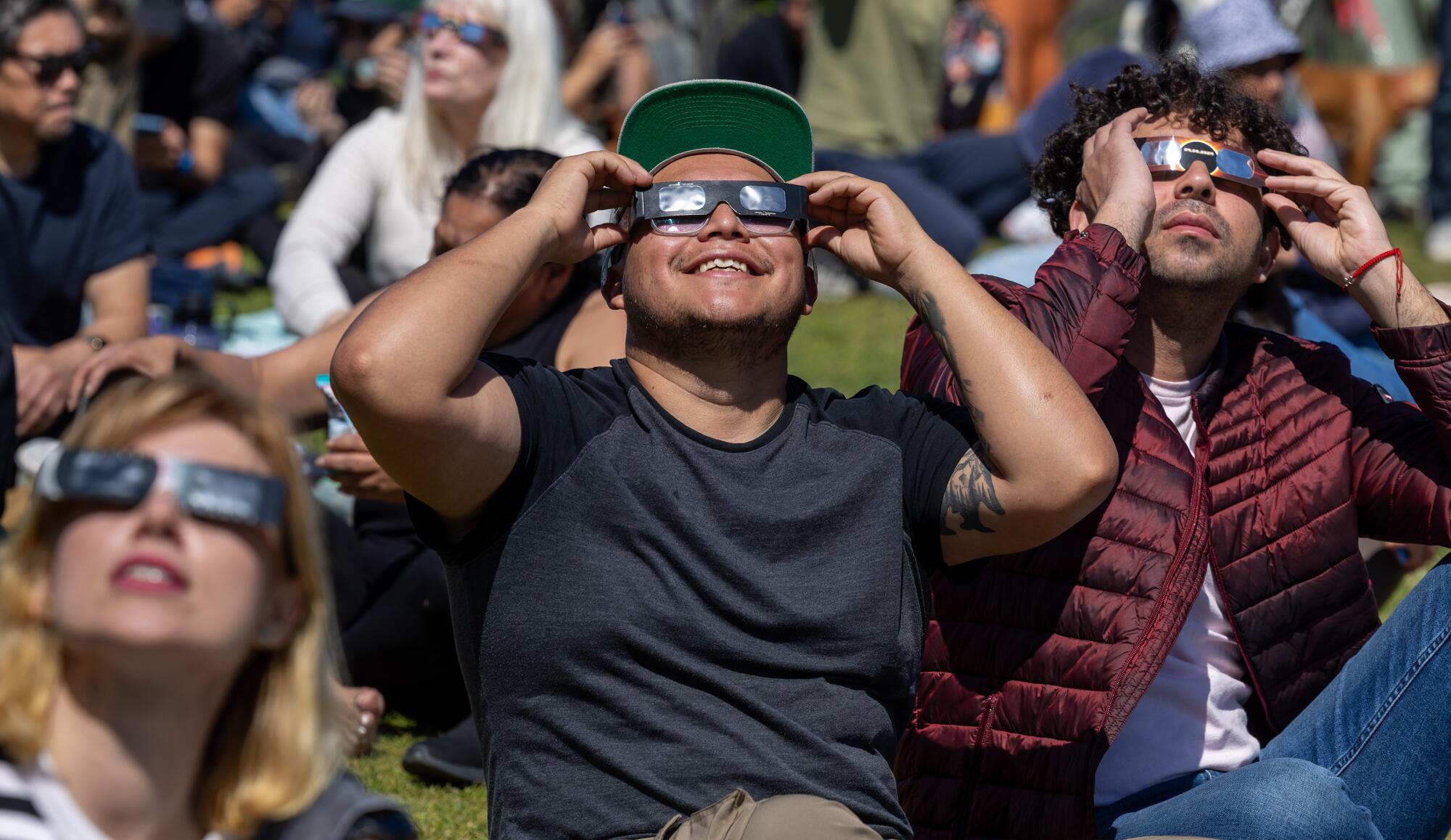 Jesus Perez sits on the grass with others in solar eclipse glasses looking up.