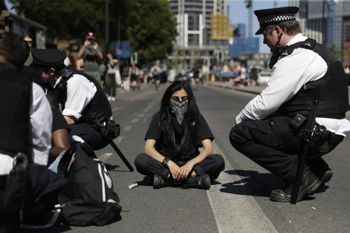 A masked woman sits in the middle of a road, flanked by British police officers in uniform 