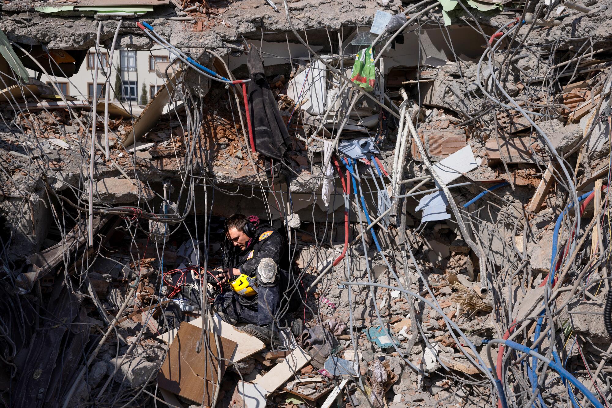 A man, wearing headphones and holding a yellow helmet, sits in a nest of rubble, which fills the whole frame.