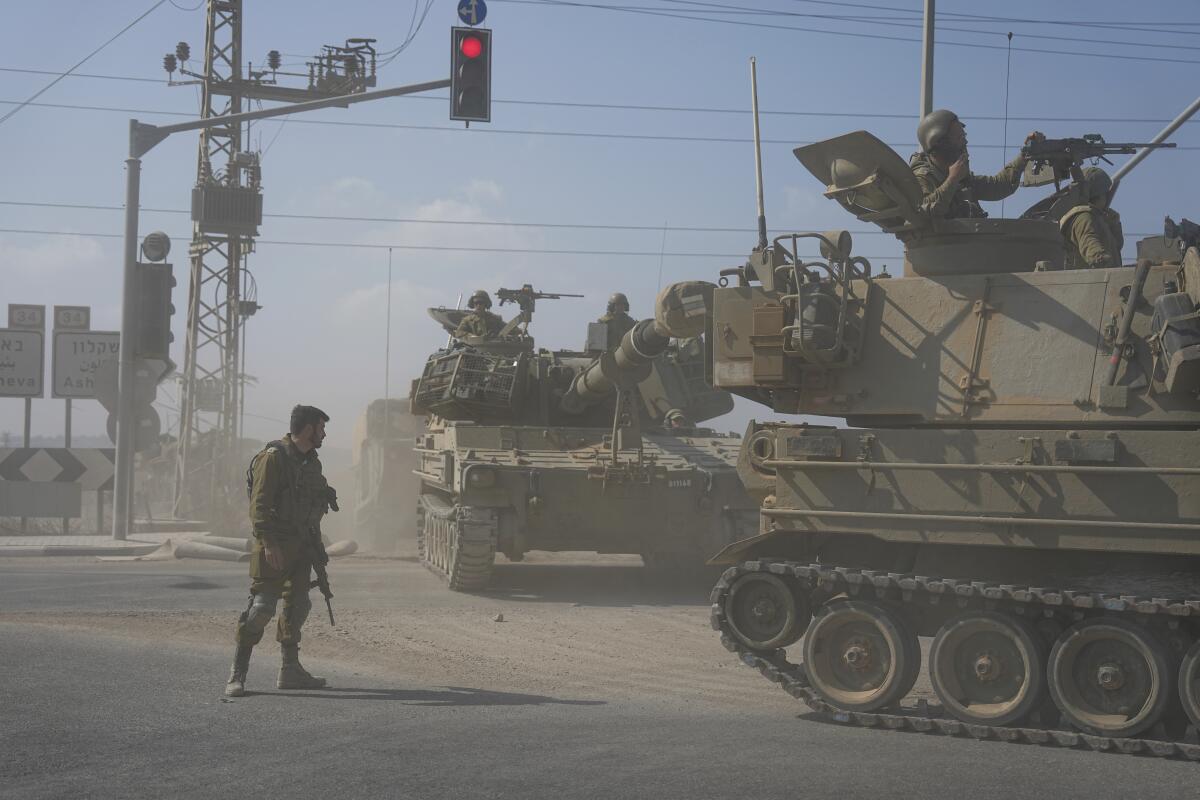 A soldier with a rifle stands by as two tanks with armed troops on top drive along a dusty street