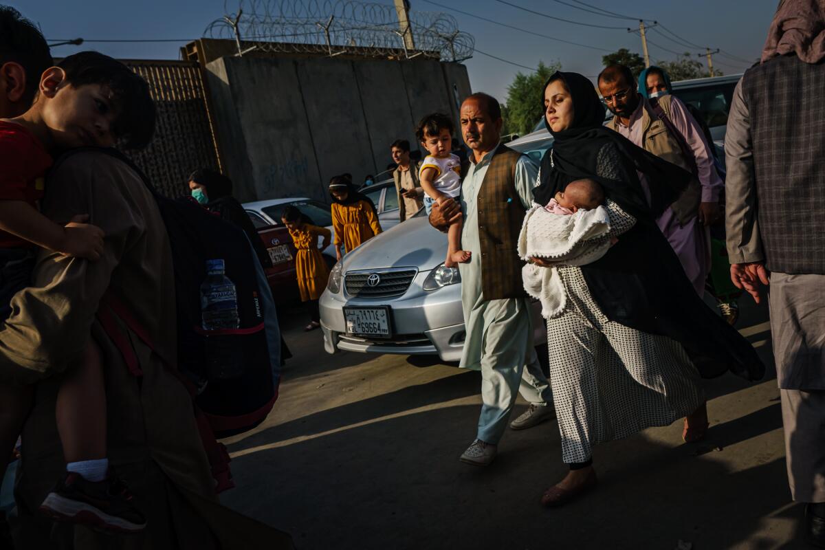 A man and a woman holding small children walk among cars and people