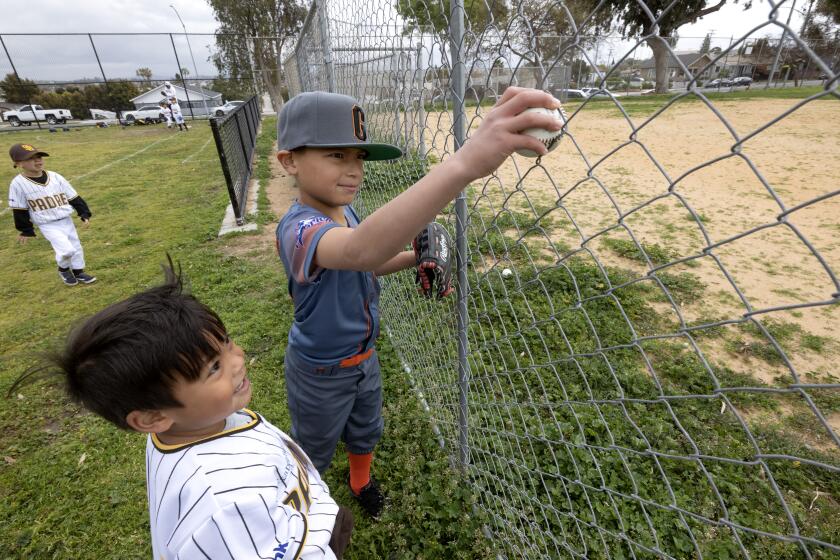 SAN DIEGO, CA - MARCH 4, 2023: Little league baseball player Benjamin Buenrostro, 7, grabs a baseball stuck in a fence surrounding a closed baseball field, right, as Logan Cervantes, 6, watches during opening day for the San Diego American Little League at Memorial Park in Logan Heights, San Diego on Saturday, March 4, 2023. The Lil Friars tee ball team would have normally practiced in the field at right, but because it's closed, have to practice at the grassy field at left. (Hayne Palmour IV / For The San Diego Union-Tribune)